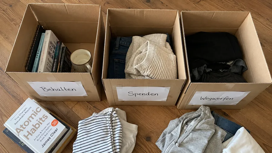 Draufsicht auf drei offene Kartons auf Holzboden mit handbeschrifteten Zetteln für Behalten, Spenden und Wegwerfen