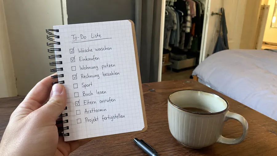 Hand hält ein kleines Notizbuch mit handgeschriebener Checkliste, daneben eine Kaffeetasse, im Hintergrund ein geöffneter Kleiderschrank