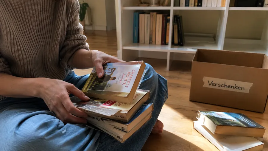 Person sitzt vor einem Bücherregal und sortiert einen Stapel gebrauchter Bücher, im Hintergrund eine Kiste mit der Aufschrift Verschenken