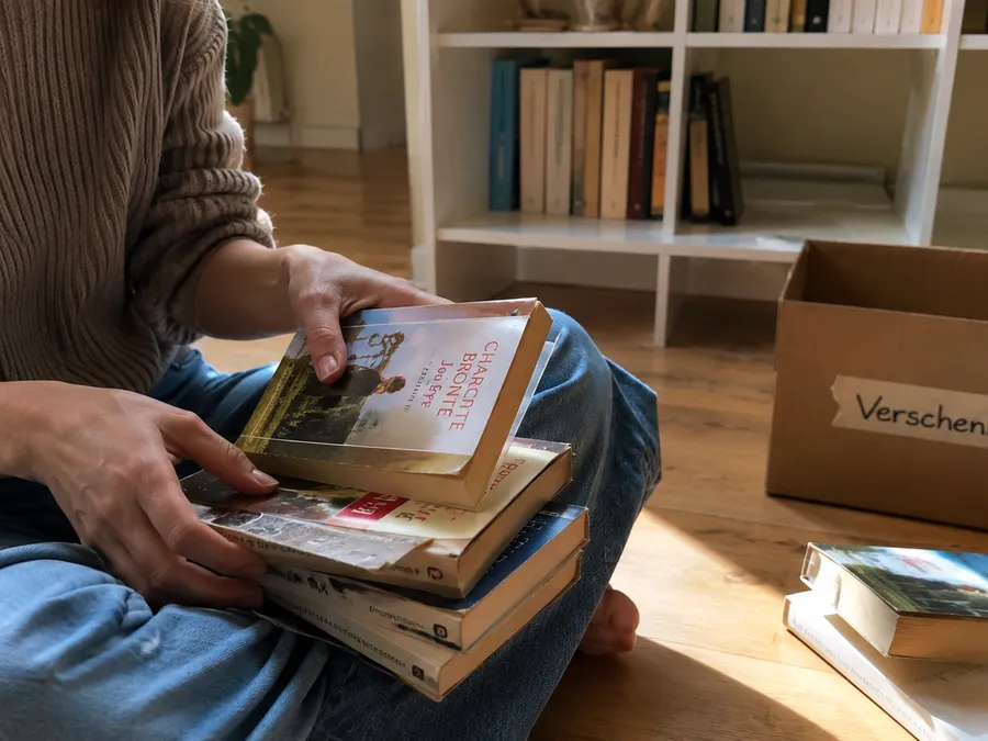 Person sitzt vor einem Bücherregal und sortiert einen Stapel gebrauchter Bücher, im Hintergrund eine Kiste mit der Aufschrift Verschenken