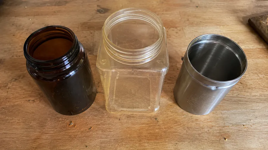 Flatlay auf Holzarbeitsplatte mit drei leeren Gewürzbehältern nebeneinander: dunkles Glas, Kunststoff, Edelstahl