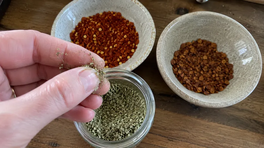 Nahaufnahme einer Hand, die eine Gewürzprobe zwischen den Fingern reibt, daneben zwei Schälchen Chiliflocken im Vergleich frisch und alt