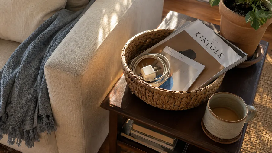 Kleiner geflochtener Korb auf einem Beistelltisch neben dem Sofa mit einem Ladekabel und Zeitschriften, daneben eine halbvolle Kaffeetasse