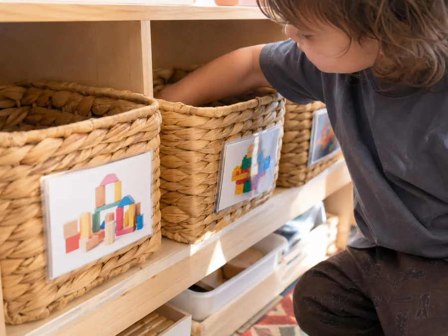 Niedriges Regal mit offenen, bildbeschrifteten Boxen in Kinderhöhe, Kind greift selbstständig hinein