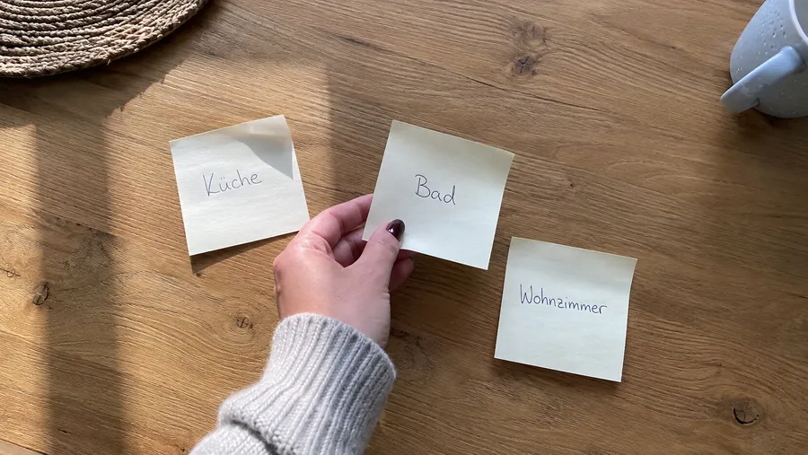 Holztisch mit drei losen Klebezetteln, beschriftet mit Küche, Bad und Wohnzimmer, eine Hand hebt unentschlossen einen Zettel hoch