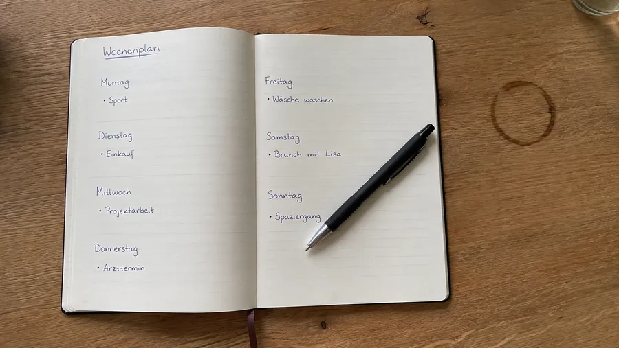 Aufsicht auf einen offenen Papierplaner mit handschriftlichen Aufgaben für jeden Wochentag, ein Stift liegt quer über der Seite